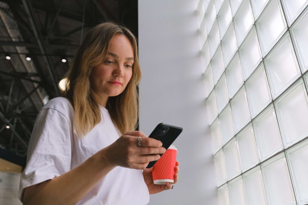 A low-angle shot of a young woman with blonde hair, wearing a white t-shirt, looking down at her black smartphone while holding a red takeaway coffee cup. She is standing next to a large wall made of glass bricks.