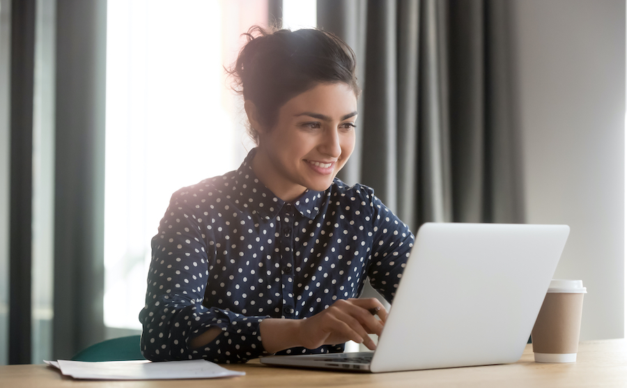 A smiling woman with her hair in a bun, wearing a dark blue polka-dot shirt, sits at a wooden desk typing on a silver laptop next to a takeaway coffee cup.